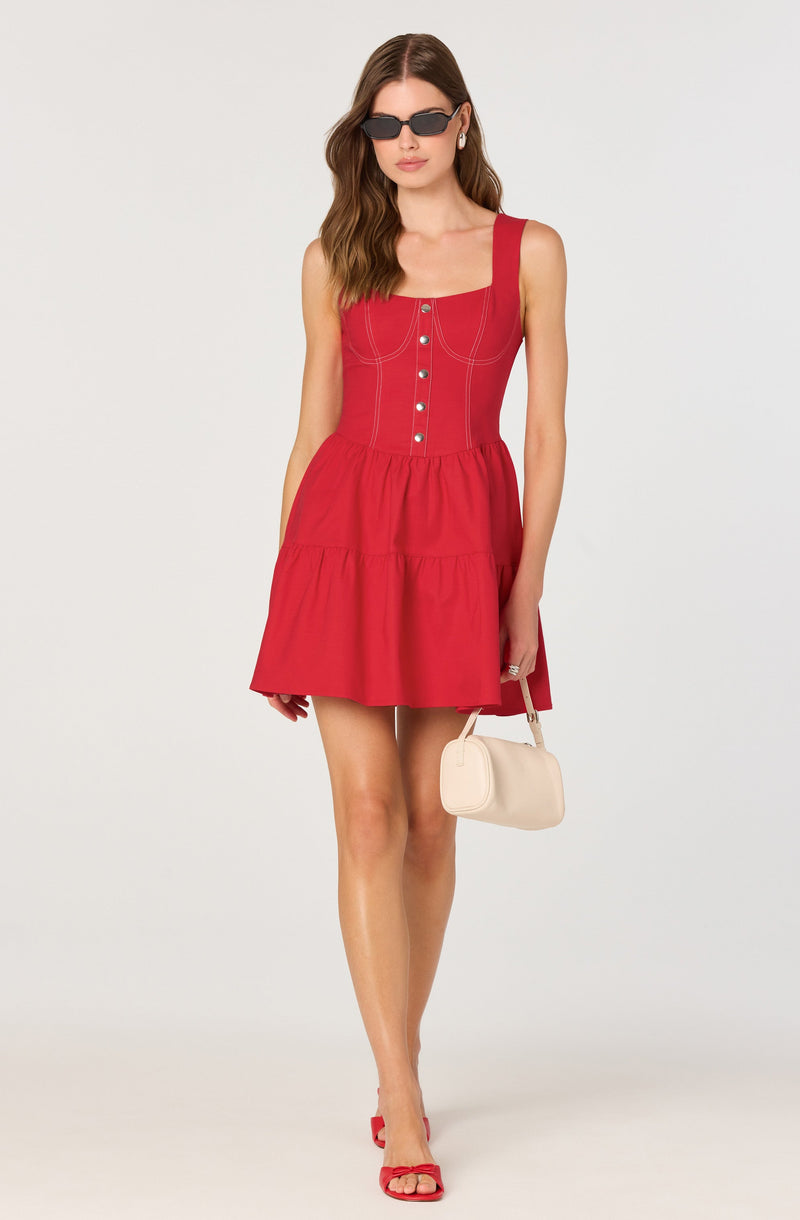 A woman is wearing a sleeveless, red dress with wide straps and a fitted bodice featuring decorative stitching and buttons down the front. The dress has a gathered waist and a tiered, flared skirt that falls above the knees. She is accessorizing with dark sunglasses, pearl earrings, and red heeled sandals with bows. She carries a small, light-colored handbag in her right hand.