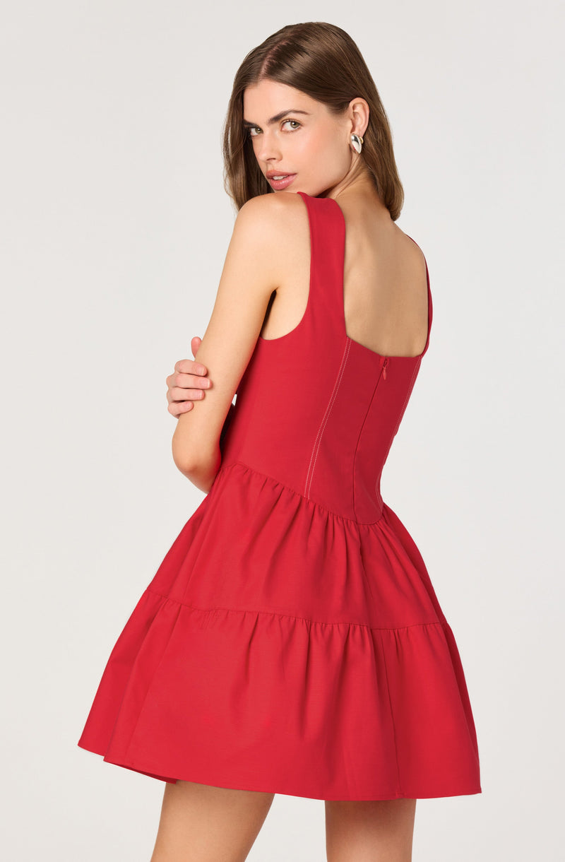 A woman is wearing a sleeveless red dress with a fitted bodice and a full, tiered skirt. The dress features wide shoulder straps and visible seams running down the back. She has earrings and is posing with her arms crossed, looking over her shoulder.