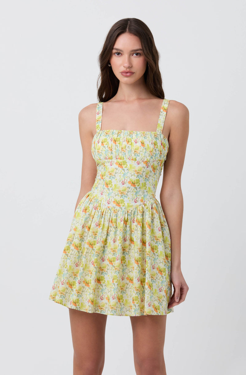 A young woman is wearing a sleeveless, knee-length dress with shoulder straps. The dress features a light, pastel-colored floral print with shades of yellow, pink, green, and blue. The bodice has pleats, and the skirt is gathered, creating a slightly flared silhouette.