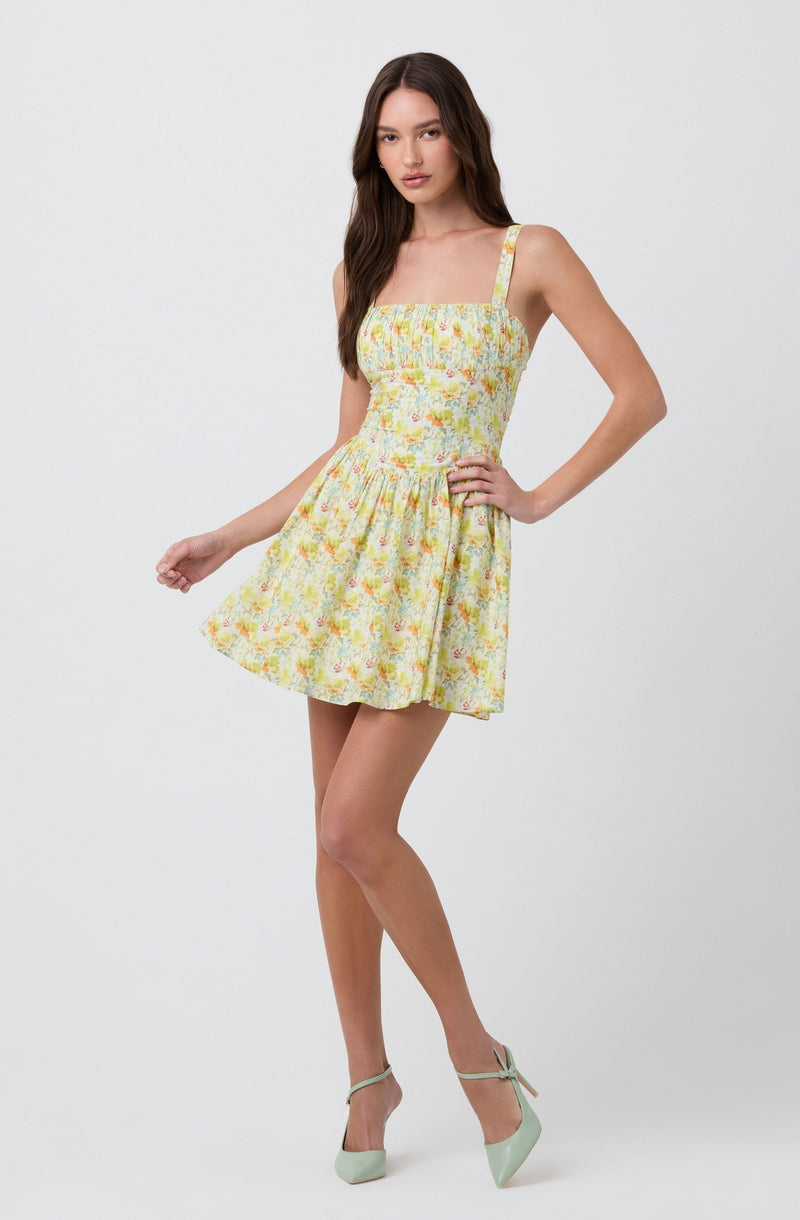 A woman is wearing a sleeveless, fitted dress with a square neckline and thin shoulder straps. The dress features a yellow background with a floral pattern in shades of pink, green, and white. The skirt is gathered at the waist, creating a flared silhouette. She is also wearing light green high-heeled shoes with ankle straps and a bow detail.