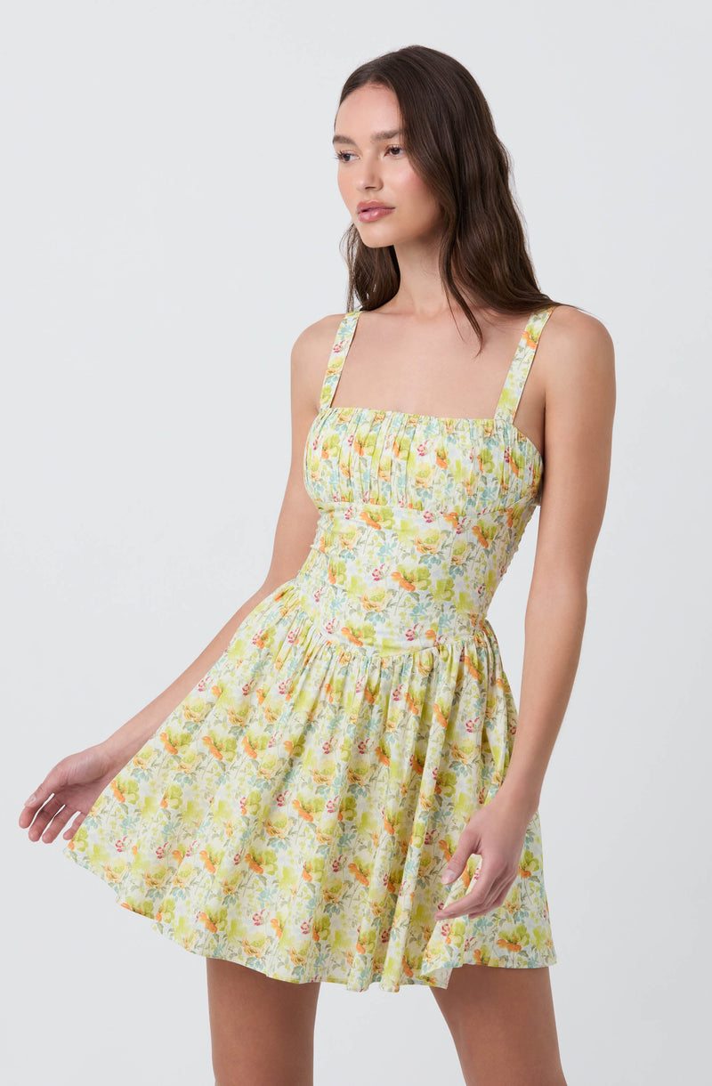 A young woman is wearing a sleeveless, knee-length dress with wide shoulder straps. The dress features a fitted bodice with gathered fabric and a floral print in soft pastel colors, including yellow, orange, pink, and green. The skirt is full and slightly flared.
