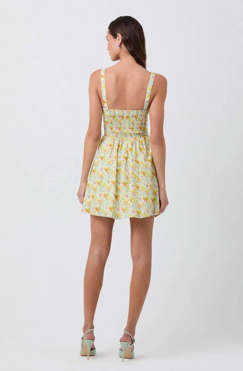A woman is wearing a sleeveless dress with wide straps. The dress features a pastel floral pattern in shades of yellow, orange, and green. It has a fitted, smocked bodice and a high waistline, with the skirt flaring out slightly to a short length above the knees. She is also wearing high-heeled sandals in a light color.