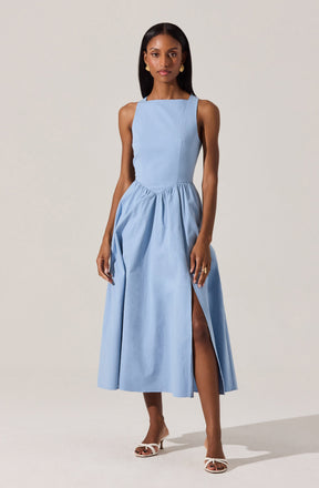 A woman is wearing a sleeveless, light blue dress with a fitted bodice and a full, gathered skirt that reaches mid-calf length. The dress features a high slit on one side, revealing her leg. She is also wearing white high-heeled sandals.