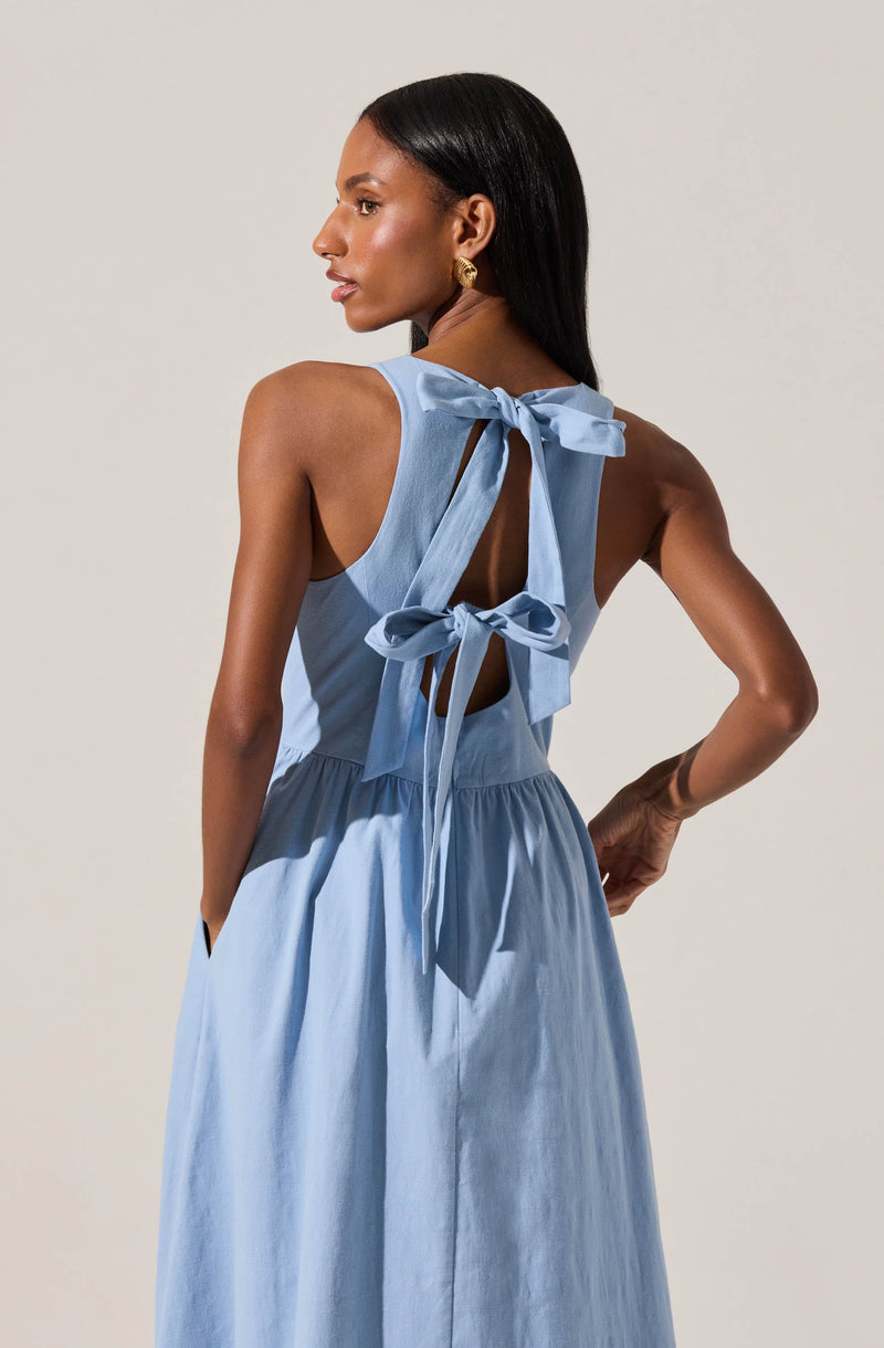 A woman is wearing a sleeveless, light blue dress with a gathered waist. The dress features a large bow at the back with two long ribbons tied into bows, and has an open back detail. She has straight, shoulder-length black hair and is accessorized with gold earrings.