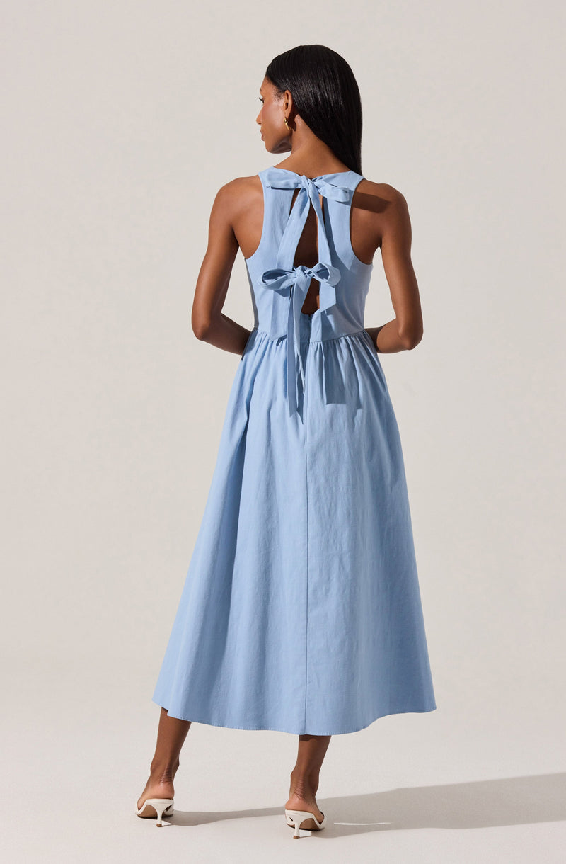 A woman is wearing a sleeveless, light blue dress with a fitted bodice that features a large bow at the back and a keyhole opening below the bow. The dress has a full, flowing skirt that reaches down to her ankles. She is also wearing white high-heeled shoes.