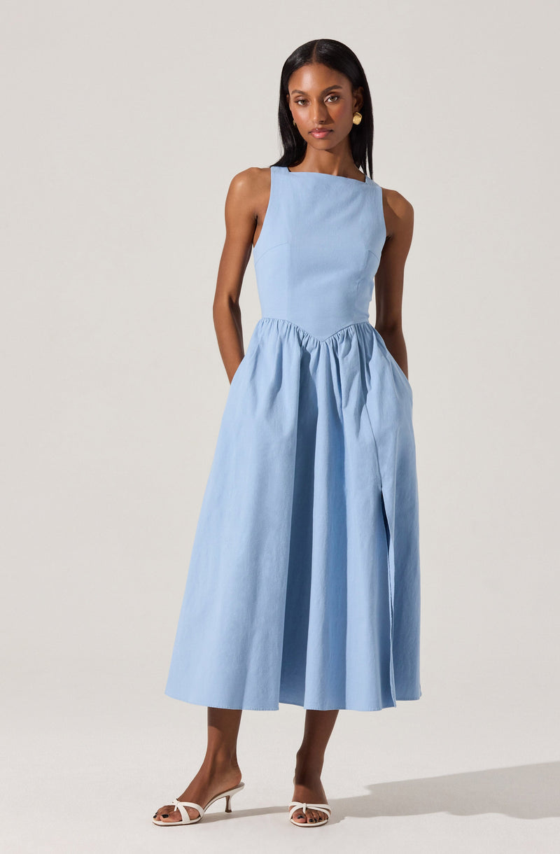 A woman is wearing a sleeveless, light blue dress with a fitted bodice and a high neckline. The dress has a gathered, full-length skirt that reaches mid-calf. She is also wearing white open-toe high-heeled sandals and earrings.