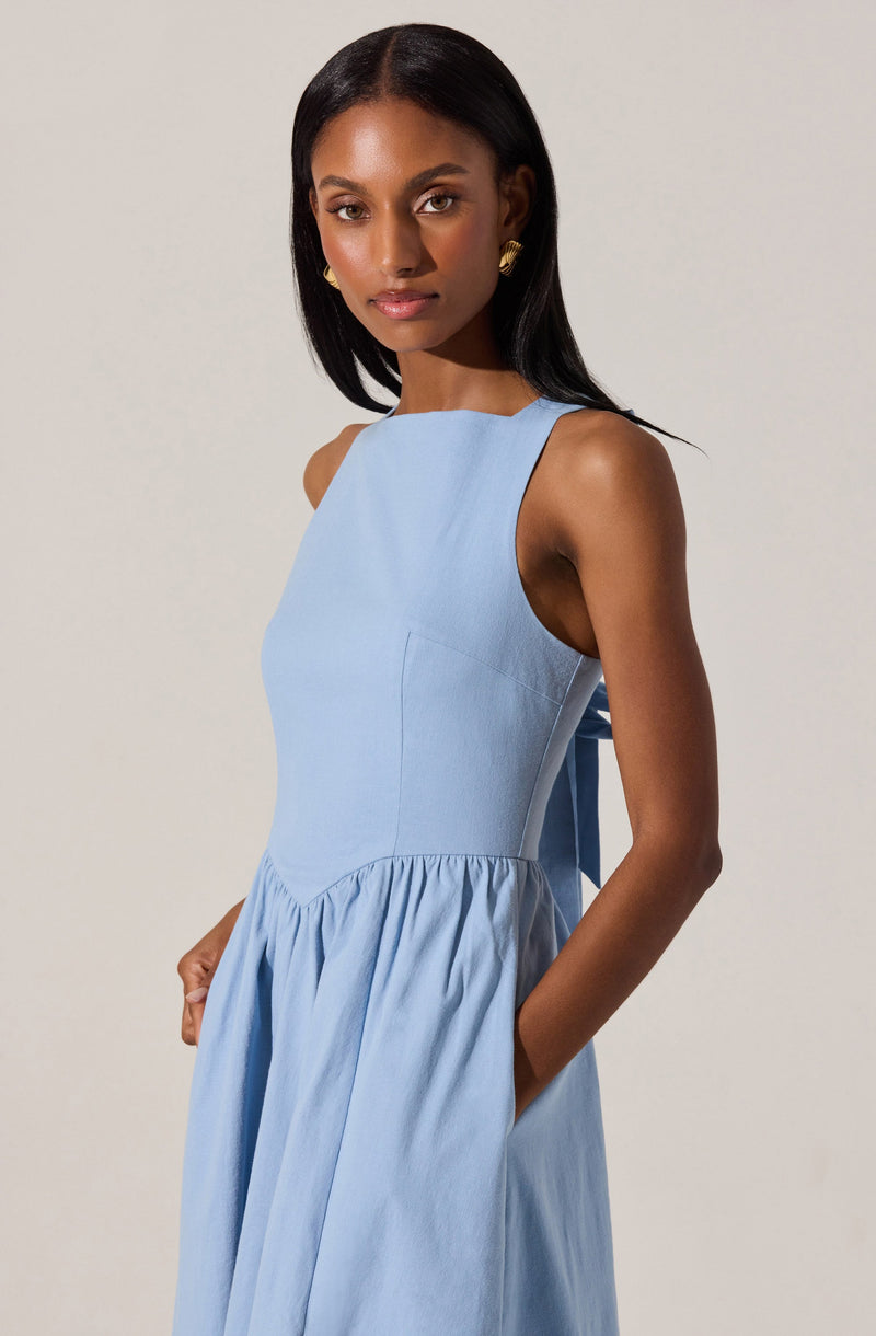 A woman is wearing a sleeveless, light blue dress with a fitted bodice and a gathered skirt. The dress features wide shoulder straps and a slightly high neckline. She has her hands in the pockets of the dress.