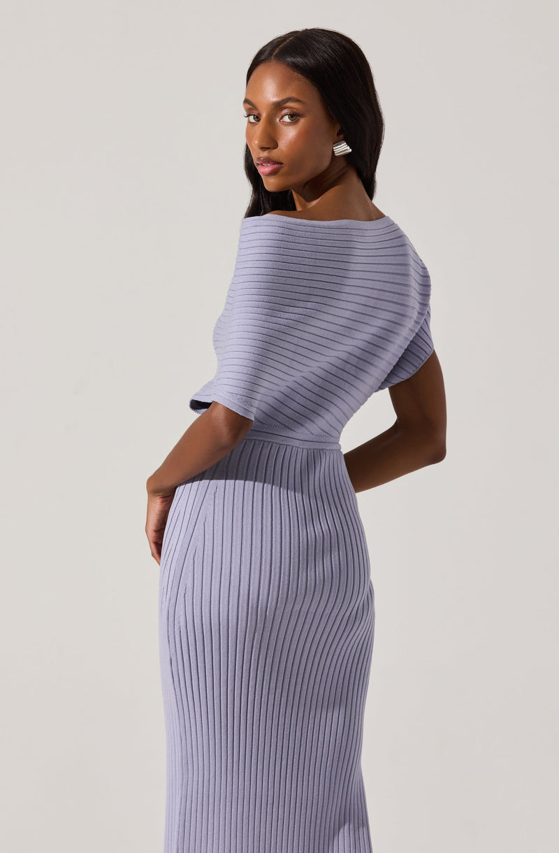 A woman is wearing a matching lilac knit dress with a ribbed pattern. The top has a loose, wide neckline and elbow-length sleeves, while the fitted skirt extends to the floor. She accessorizes with rectangular earrings and has straight, dark hair.