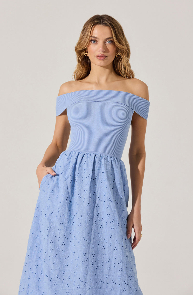 A woman is wearing an off-the-shoulder dress with a fitted, ribbed bodice in a light blue color. The skirt is made of a slightly darker blue fabric with an eyelet pattern, creating a textured and airy appearance. The dress has a relaxed and elegant style, and she has one hand in her pocket.