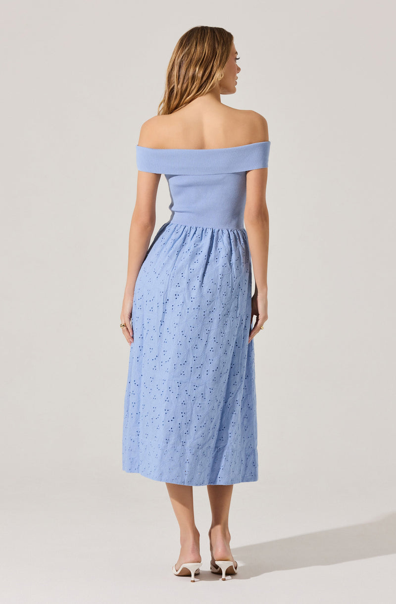 A woman is wearing an off-the-shoulder, light blue top with a wide, ribbed neckline that folds over. She has paired it with a long, flowy skirt in a matching light blue shade, featuring eyelet embroidery throughout. She is also wearing white high-heeled shoes.