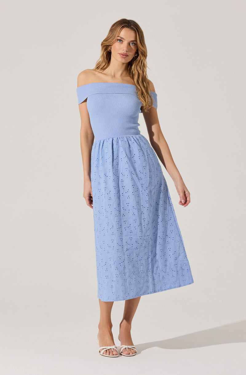 A woman is wearing an off-the-shoulder, light blue, fitted ribbed top with a wide neckline. She is also wearing a long, light blue skirt with eyelet embroidery and a gathered waistband. She has on white open-toe sandals.