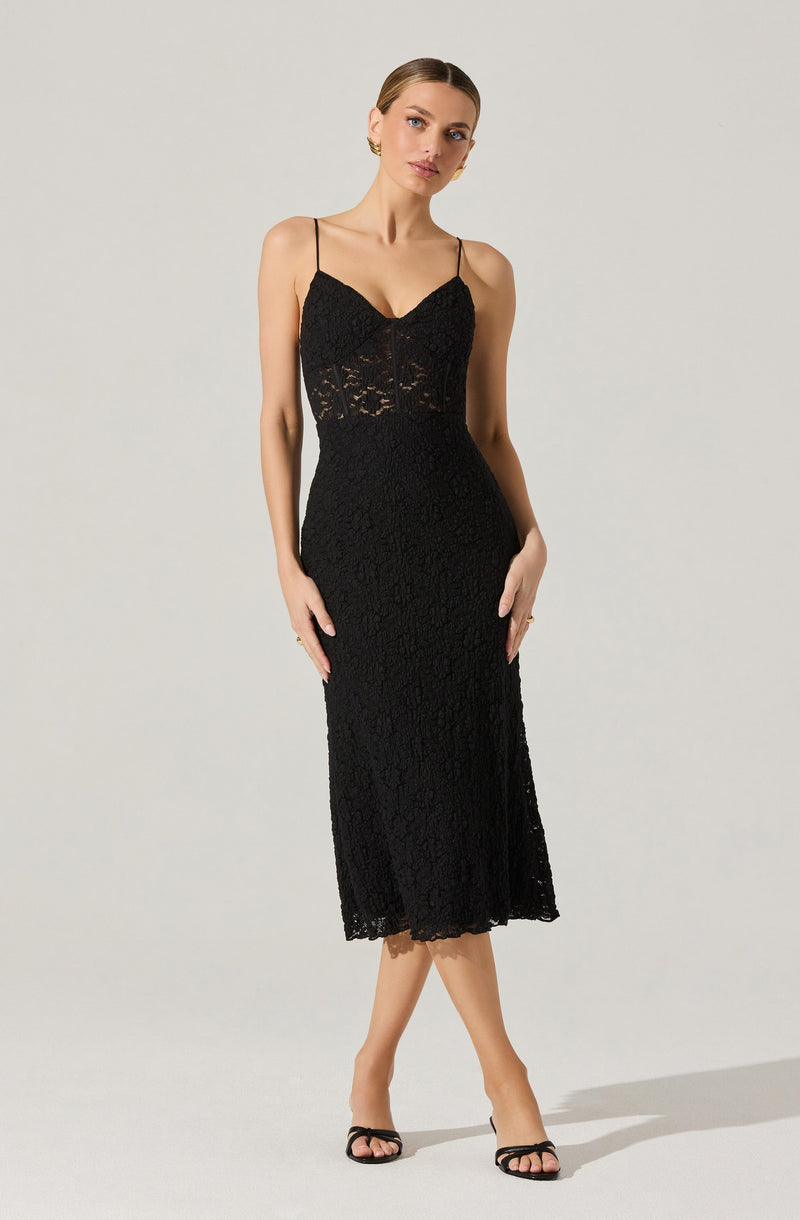 A woman is wearing a black lace midi dress with spaghetti straps, featuring a fitted bodice with a sweetheart neckline and a sheer lace panel at the waist. The skirt falls below the knees with a scalloped hem, and she pairs the dress with black open-toe heels.