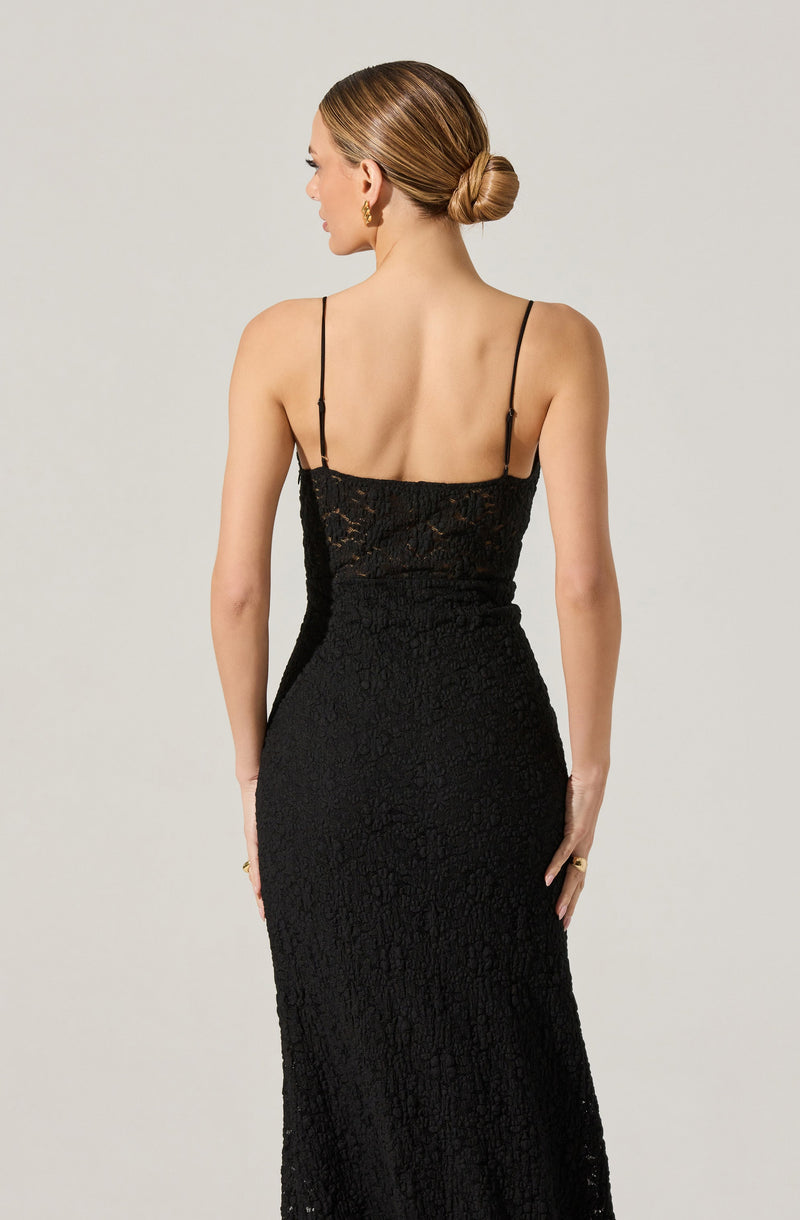 A woman is wearing a black, sleeveless, full-length dress with thin spaghetti straps. The dress features a lace overlay with a floral pattern and a fitted silhouette that accentuates her figure.