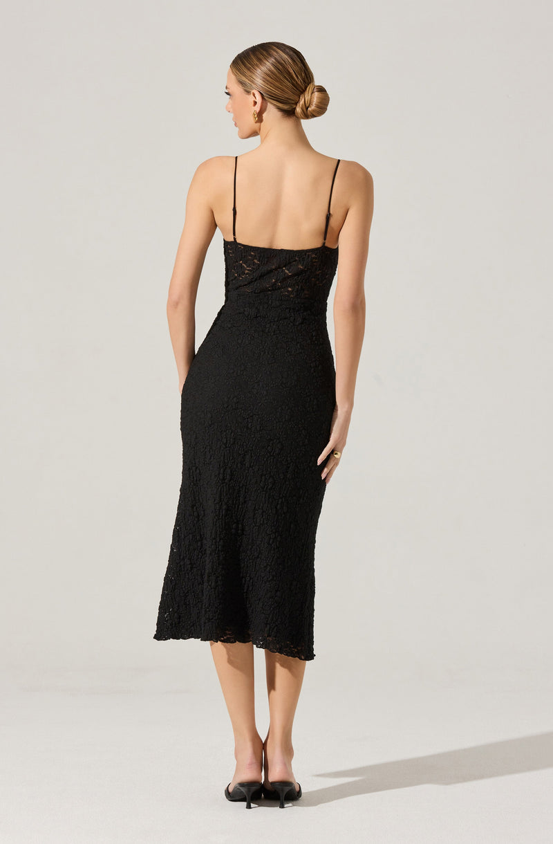 A woman is wearing a sleeveless, black lace dress with thin spaghetti straps. The dress has a fitted bodice and falls to mid-calf length with a slightly scalloped hem. The lace fabric features a floral pattern, giving it an elegant and textured appearance.