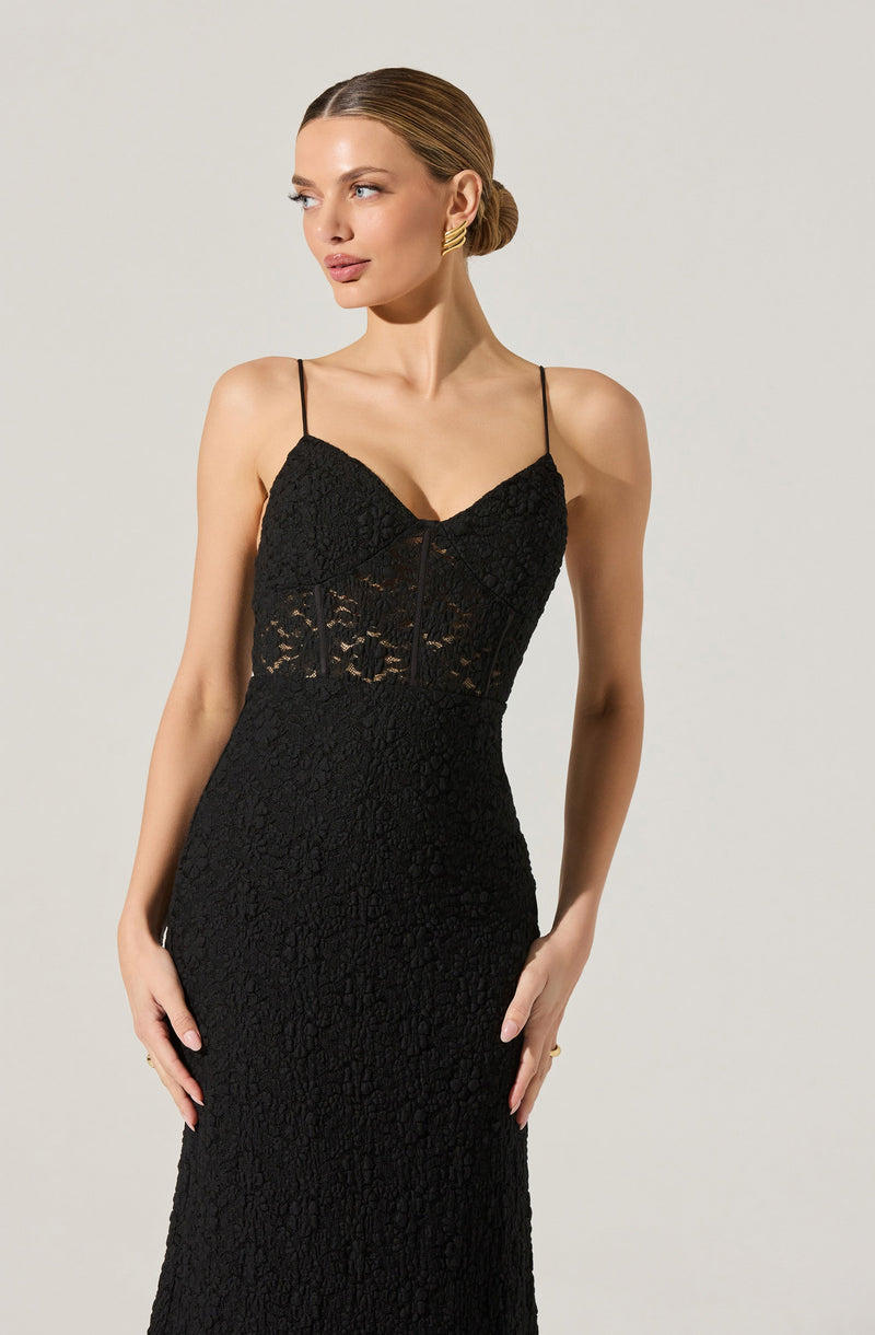 A woman is wearing a black lace dress with spaghetti straps. The dress features a fitted bodice with a lace overlay and a sheer lace panel around the midsection. The skirt is long and form-fitting, made of the same textured lace fabric throughout.
