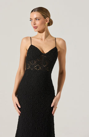A woman is wearing a black lace dress with spaghetti straps. The dress features a fitted bodice with a lace overlay and a sheer lace panel around the midsection. The skirt is long and form-fitting, made of the same textured lace fabric throughout.