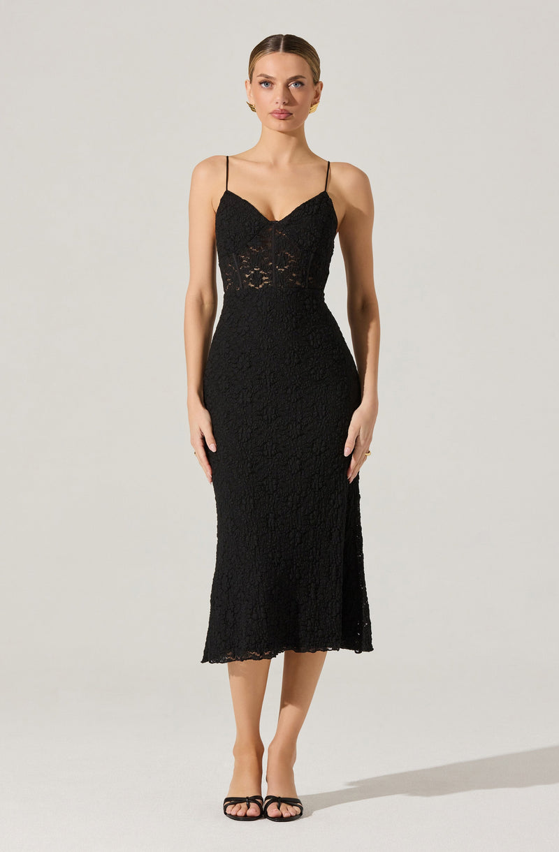 A woman is wearing a black, fitted lace dress with thin spaghetti straps. The dress features a sweetheart neckline and the lace pattern is floral and intricate throughout. The hemline of the dress falls to mid-calf length. She is also wearing black open-toed high heels.