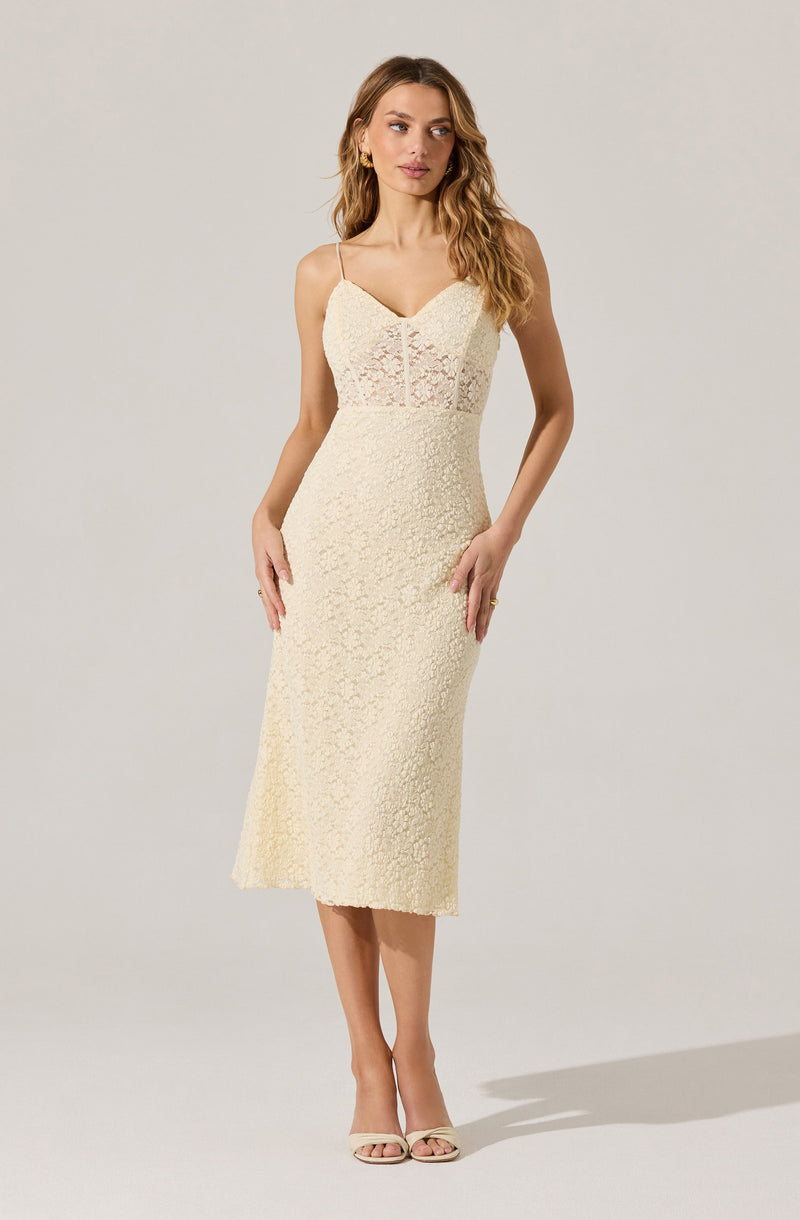 A woman is wearing a sleeveless, cream-colored, lace midi dress with thin spaghetti straps. The dress features a fitted silhouette with a V-neckline and a sheer lace panel on the upper chest and midsection. She is also wearing open-toe heels in a matching light color.