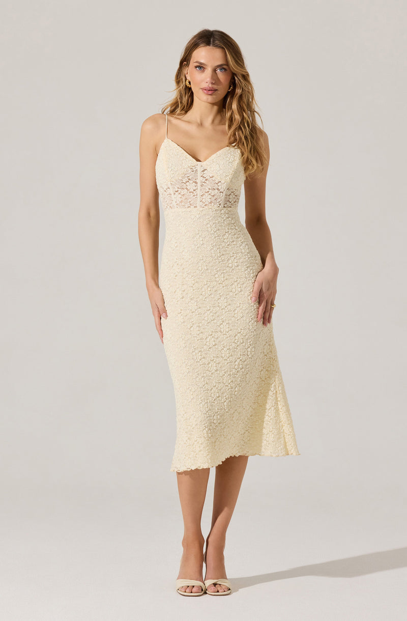 A woman is wearing a sleeveless, cream-colored dress with thin spaghetti straps. The dress has a fitted bodice with a sweetheart neckline, featuring a semi-sheer lace panel at the top. The skirt of the dress extends to mid-calf and is textured with a floral lace pattern throughout. She is also wearing open-toed, strappy high-heeled sandals in a matching light color.