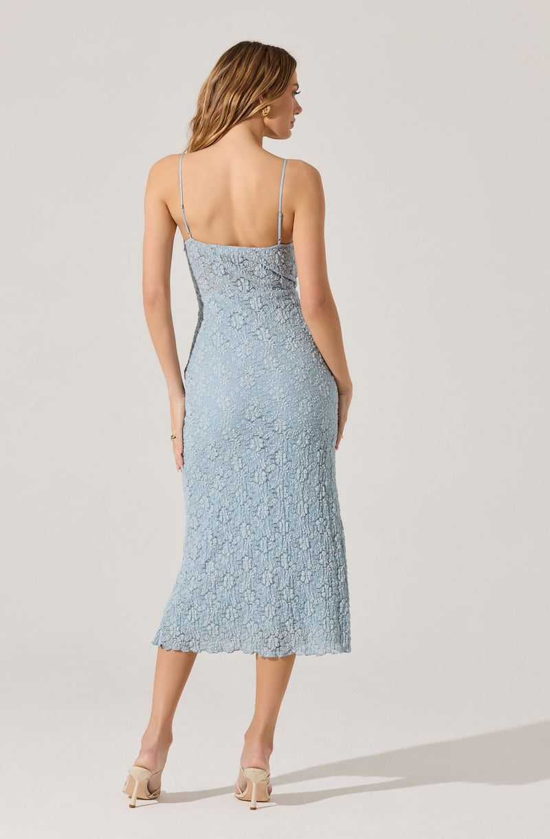 A woman is wearing a light blue, textured, midi-length dress with thin spaghetti straps. The dress has a fitted silhouette and appears to have a subtle floral or abstract pattern in the fabric. She is also wearing nude high-heeled shoes.