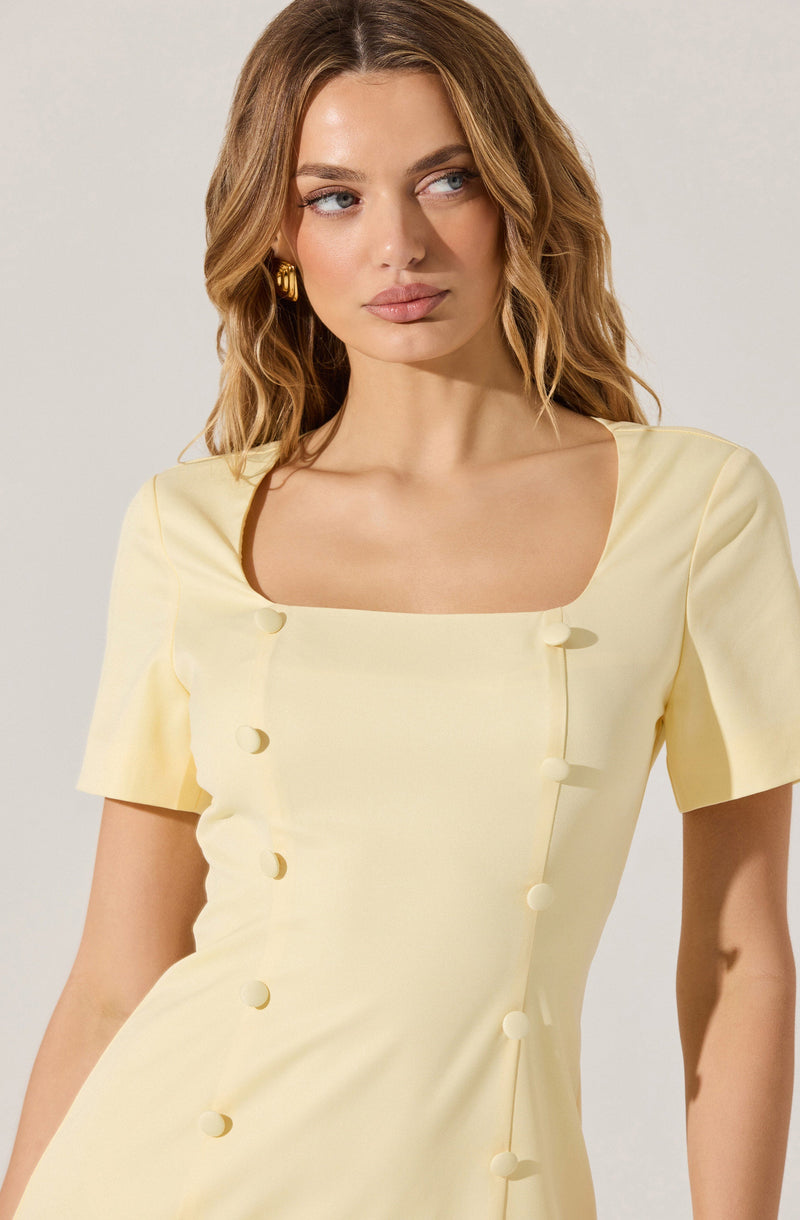 A woman is wearing a light yellow dress with a square neckline and short sleeves. The dress features large decorative buttons running diagonally along the front.