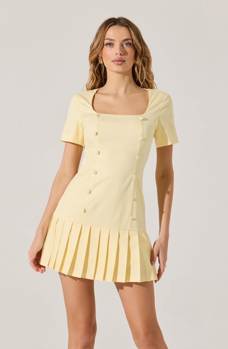 A woman is wearing a short, light yellow dress with short sleeves. The dress features a square neckline with brown piping around the edges, a row of fabric button details down the front, and a pleated skirt with wide, evenly spaced pleats at the hem.