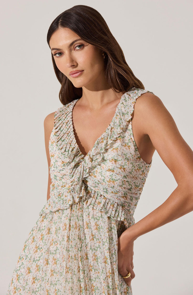 A woman is wearing a sleeveless dress with a floral print featuring shades of green, yellow, and pink. The dress has ruffled details along the V-neckline and armholes, with a ruffled hem at the waist. It is gathered at the waist with a fabric tie, creating a relaxed and feminine look.