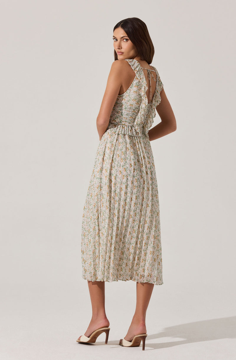A woman is wearing a sleeveless, floral-patterned dress with ruffles on the shoulder straps and back. The dress has a fitted bodice with a keyhole opening at the back and an elastic waistband, flowing into a pleated, ankle-length skirt. She is also wearing high-heeled sandals with a wide strap across the toes and a thin ankle strap.