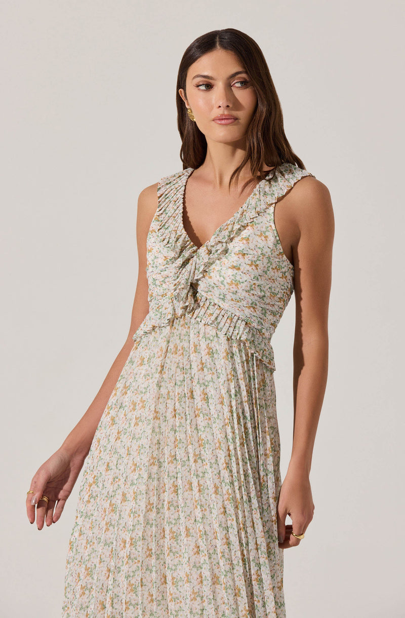 A woman is wearing a sleeveless, V-neck dress with ruffled trim along the neckline and shoulders. The dress features a floral pattern with small, multicolored flowers and has pleated details on the skirt, which include vertical stripes in coordinating colors.