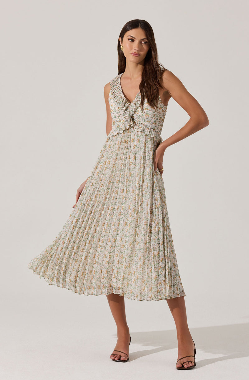 A woman is wearing a sleeveless, V-neck, midi-length dress with a floral print pattern. The dress has a ruffled detail at the neckline and waistband, and features a pleated skirt. She is also wearing open-toed, high-heeled sandals.