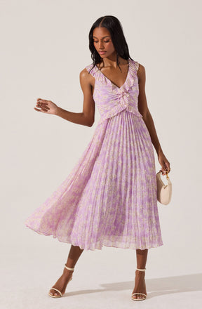 A woman is wearing a sleeveless, light purple dress with a V-neckline and ruffle details around the shoulders and neckline. The dress features a fitted bodice and a full, pleated skirt with a subtle floral pattern in pastel shades. She holds a small beige handbag in her right hand and is wearing light-colored, open-toe high heel sandals.