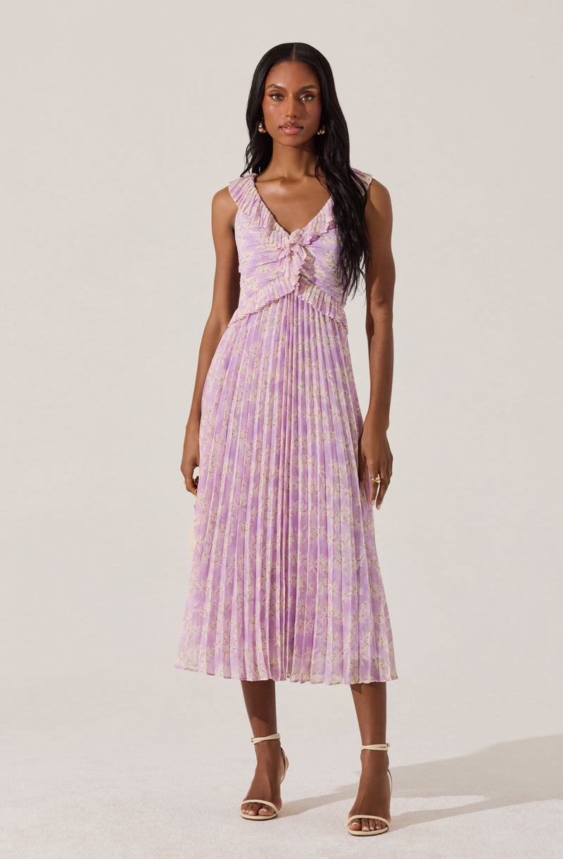 A woman is wearing a sleeveless, V-neck dress with a fitted bodice and a pleated skirt. The dress features a pastel pink and lavender floral pattern, with a ruffle detail at the neckline and a fabric bow at the waist. She is also wearing open-toe high heel sandals in a nude color.