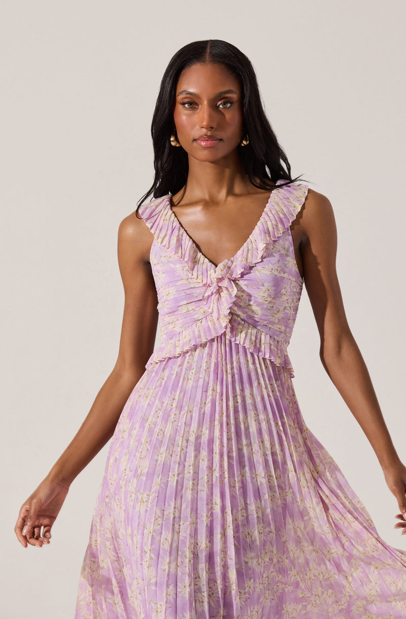 A woman is wearing a sleeveless, ankle-length dress with a V-neckline and ruffled trim. The dress is made of a pleated fabric with a light pink and lavender tie-dye or floral pattern. The bodice features a ruffled detail that cascades from the shoulders down the front.