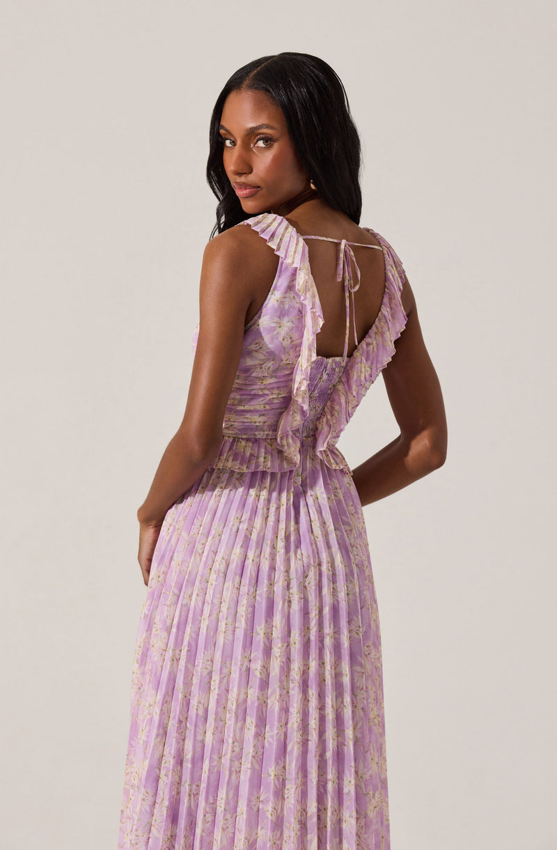 A woman is wearing a sleeveless, lavender-colored dress with a floral pattern. The dress features ruffled details on the shoulder straps and along the back opening, which is tied with a small bow at the top. The skirt of the dress is pleated, creating a flowing and elegant silhouette.