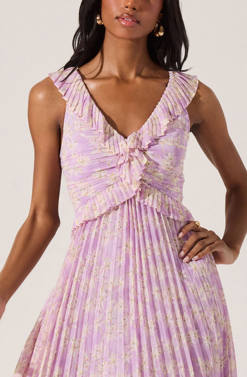 A woman is wearing a sleeveless, V-neck dress with ruffled trim along the neckline and waist. The dress has a light purple and yellow floral pattern with pleated fabric throughout.