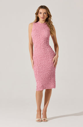 A woman is wearing a sleeveless, knee-length pink textured dress with a high neckline. She is also wearing nude-colored open-toe heels and gold earrings.