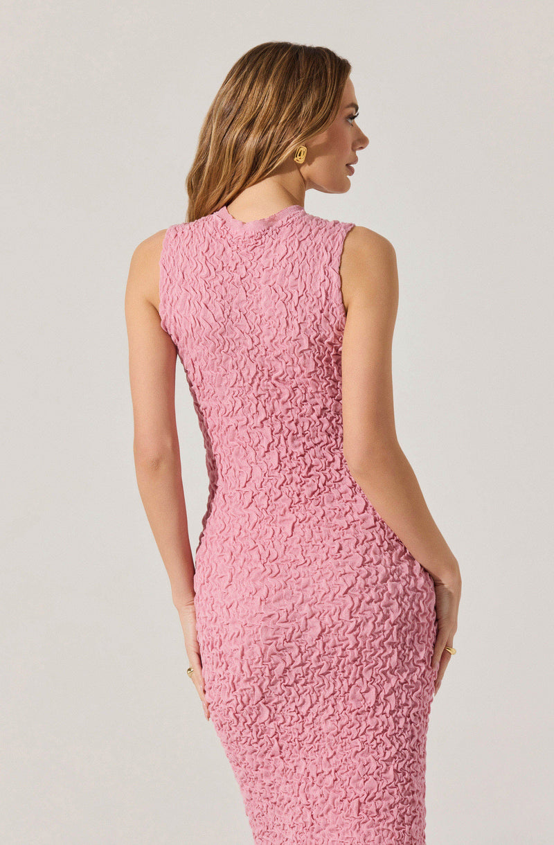 A woman is wearing a sleeveless, fitted dress in a soft pink color with a textured, ruched fabric. The dress has a high neckline and is form-fitting, accentuating her waist and hips.