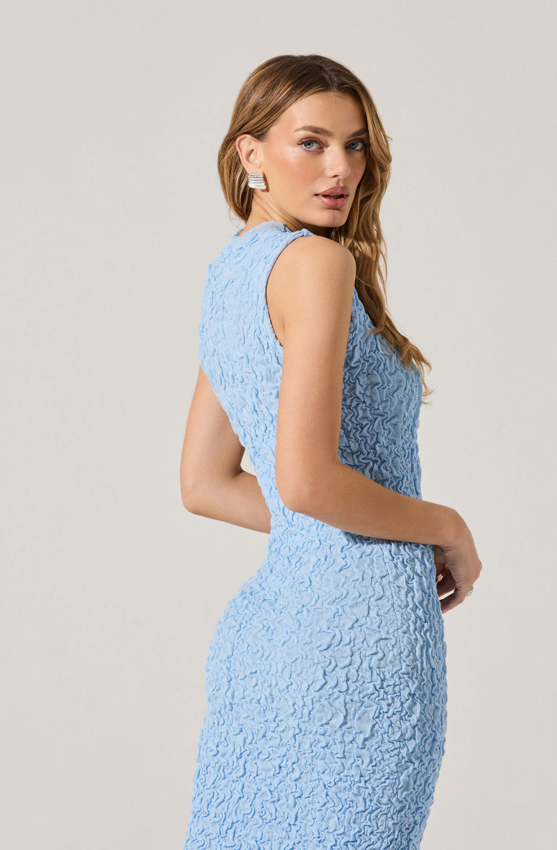 A woman is wearing a sleeveless, textured light blue dress with a fitted silhouette. The dress features a high neckline and appears to be made of a ruched or crinkled fabric. She is accessorized with large, rectangular, silver earrings and has wavy, light brown hair.
