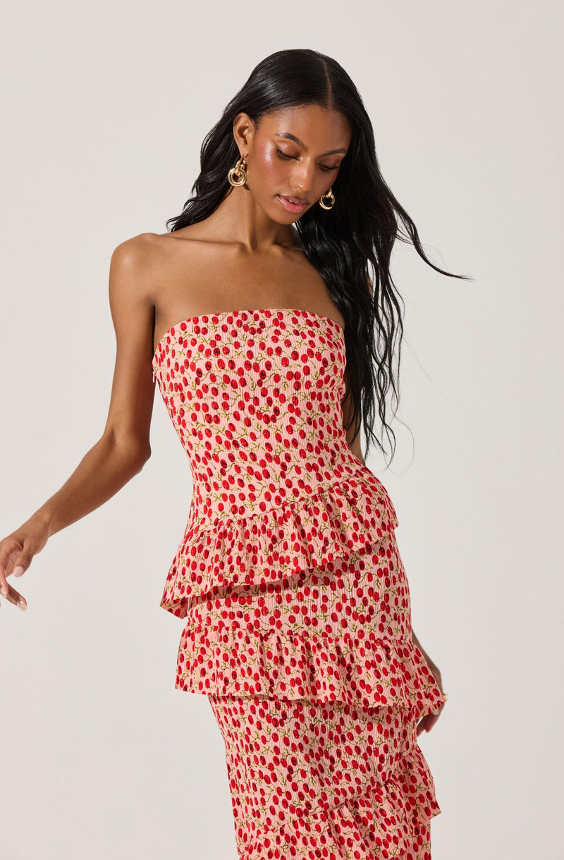 A woman is wearing a strapless, tiered dress with a pattern of red cherries and green stems on a beige background. She has long dark hair and is accessorized with large, gold hoop earrings.