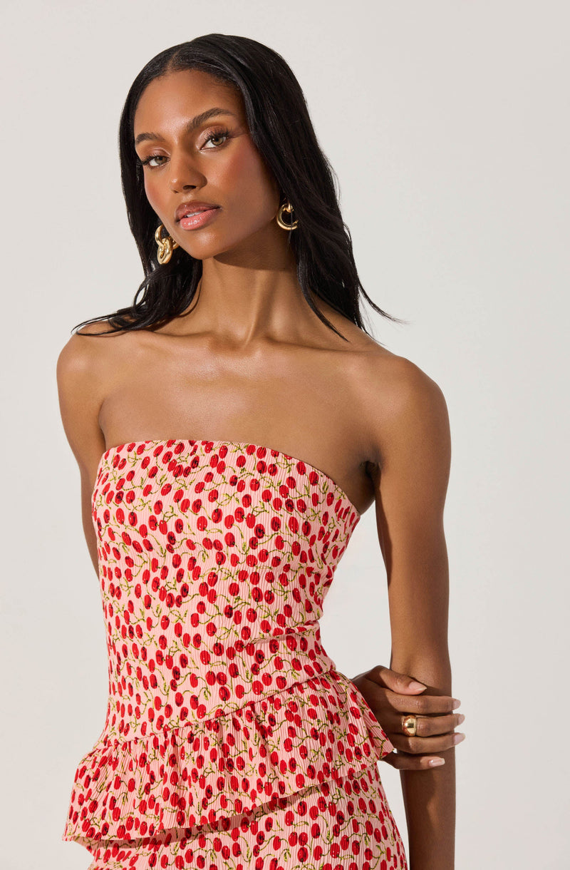 A woman is wearing a strapless dress with a fitted bodice and a peplum detail at the waist. The dress features a pattern of small red cherries with green stems on a light pink background. She accessorizes with large gold hoop earrings and a gold ring on her right hand.