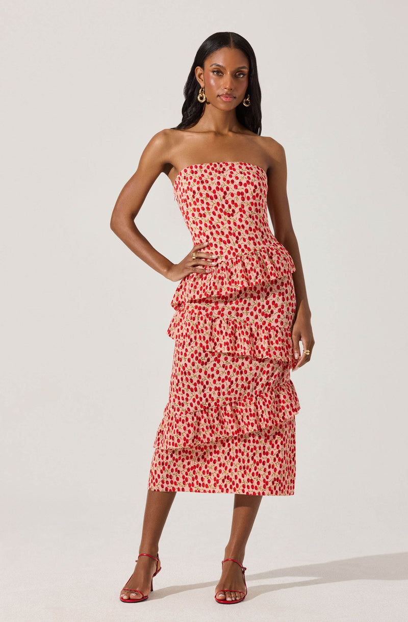 A woman is wearing a strapless, tiered midi dress with a red and beige floral pattern. She has dark, wavy hair, gold hoop earrings, and red high-heeled sandals.