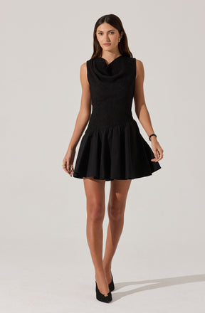 A woman is wearing a sleeveless black dress with a fitted, textured bodice and a wide, gathered skirt that flares out above the knees. The dress has a high neckline with a draped detail. She is also wearing black high-heeled shoes.