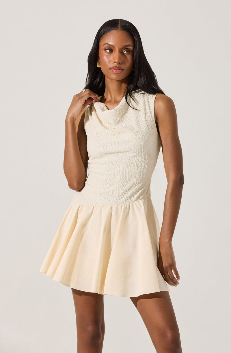 A woman is wearing a sleeveless, off-white dress with a textured, ruched bodice and a wide, draped neckline. The skirt is pleated and flares out, ending above the knees.