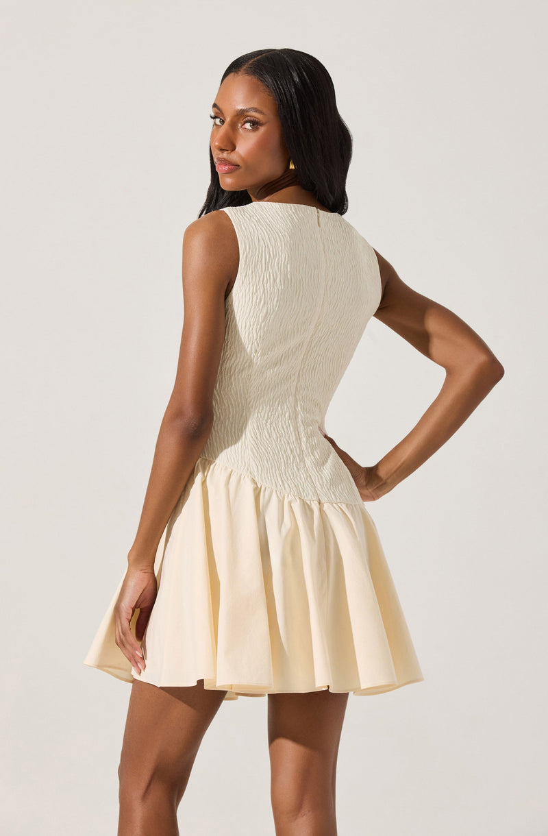 A woman is wearing a sleeveless, cream-colored dress with a fitted, textured bodice and a flared, pleated skirt. The dress has a keyhole opening at the back with a small button closure.