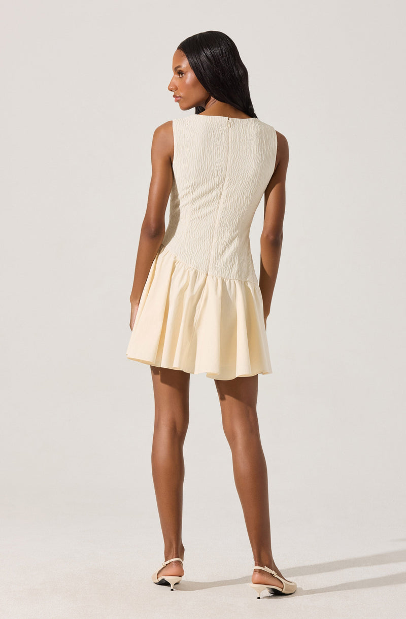A woman is wearing a sleeveless, cream-colored dress with a fitted, textured bodice and a flared, ruffled skirt. The dress has a round neckline and a hidden zipper closure at the back. She is also wearing a pair of beige, open-toe heels with ankle straps.