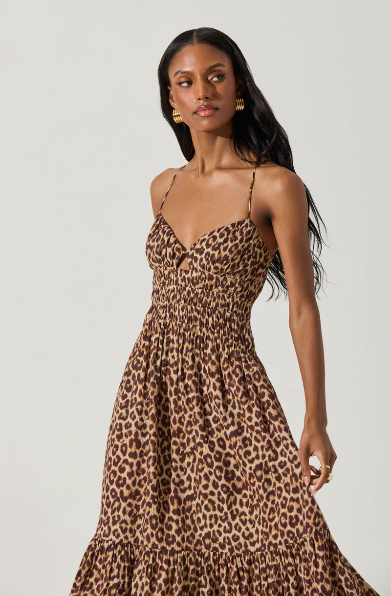 A woman is wearing a sleeveless, leopard print dress with thin spaghetti straps. The dress has a fitted bodice with a slight sweetheart neckline and an elasticated waist that creates a gathered effect. The skirt is long and flowy, featuring a tiered ruffle at the bottom.