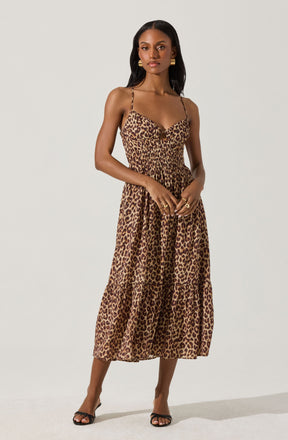 A woman is wearing a sleeveless, leopard print midi dress with thin spaghetti straps and a sweetheart neckline. The dress features a gathered waist and a tiered skirt that falls below the knees. She accessorizes with large gold hoop earrings, multiple rings, and open-toed black heeled sandals.