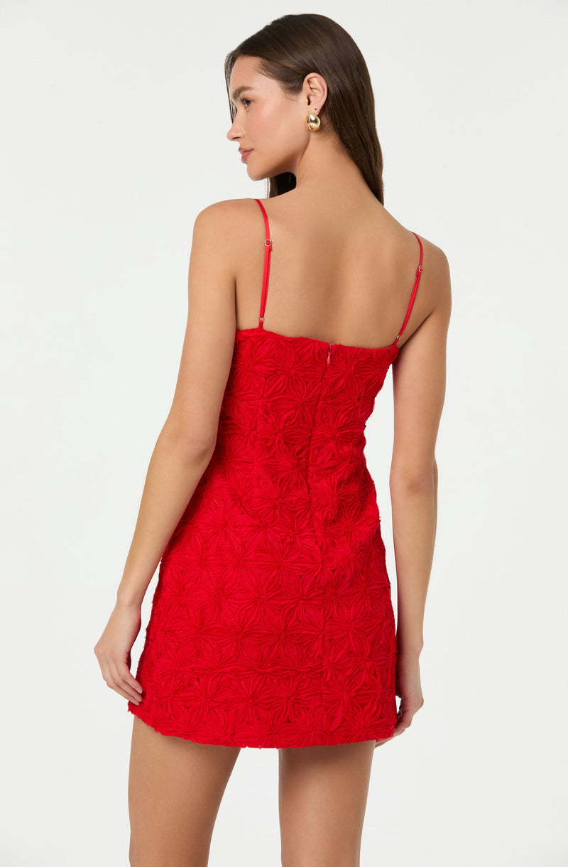 A woman is wearing a red dress with adjustable spaghetti straps. The dress features a textured floral or geometric pattern that creates a ruched effect all over the fabric. The length is above the knee, and the fit is form-fitting. She is accessorizing with large gold hoop earrings.
