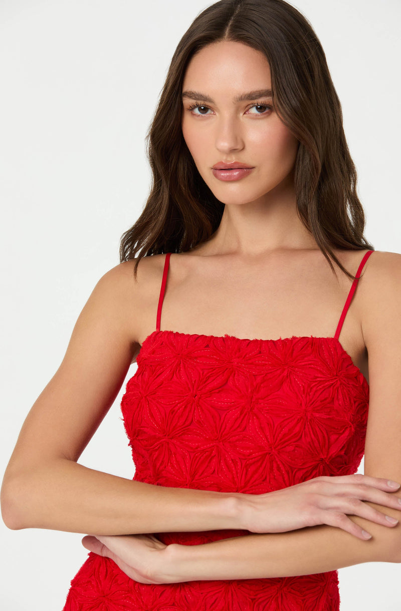 A woman is wearing a red sleeveless dress with thin spaghetti straps. The dress features an intricate textured floral pattern throughout.