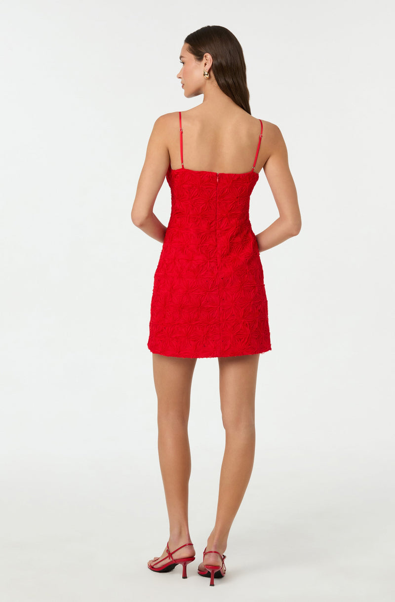 A woman is wearing a sleeveless red dress with thin spaghetti straps. The dress features a textured, floral-like pattern throughout. She is also wearing red heeled sandals with thin straps. Her hair is long and straight, and she has gold hoop earrings. She is standing against a plain, light-colored background.